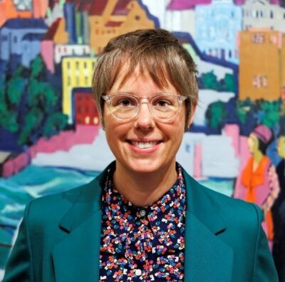 Melissa wearing a floral shirt and blue blazer, smiling in front of a patterned wall.