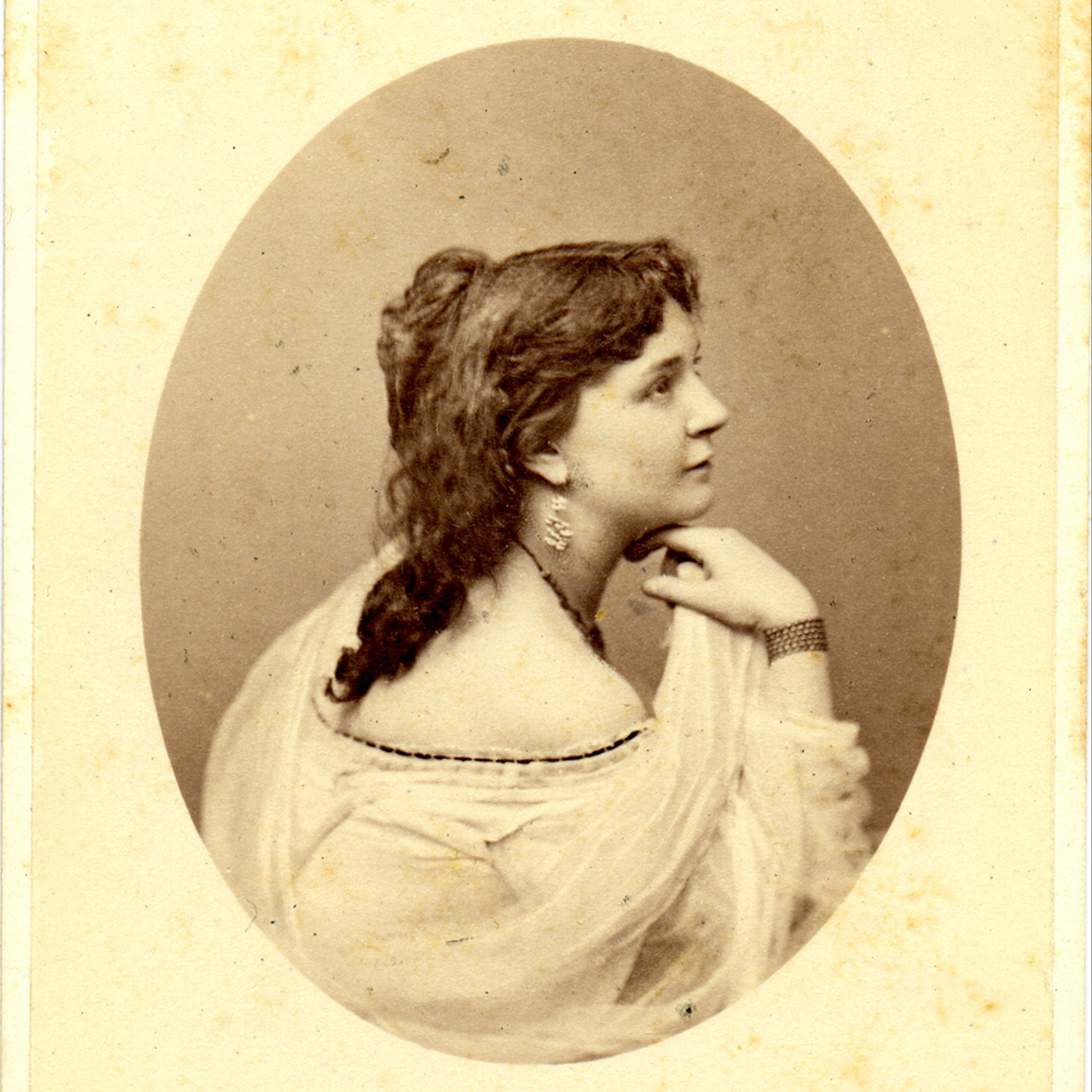 A 19th-century photograph of a woman with long hair, showing her face in profile and a hand on her chin, with her back turned to towards the camera.