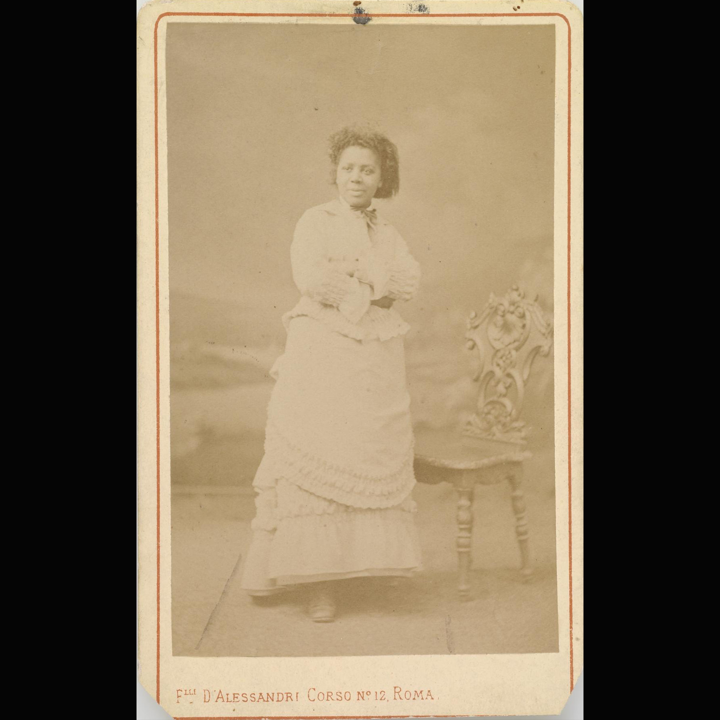 Archival photograph of Black woman standing, facing the camera, with her arms crossed and a chair behind her.
