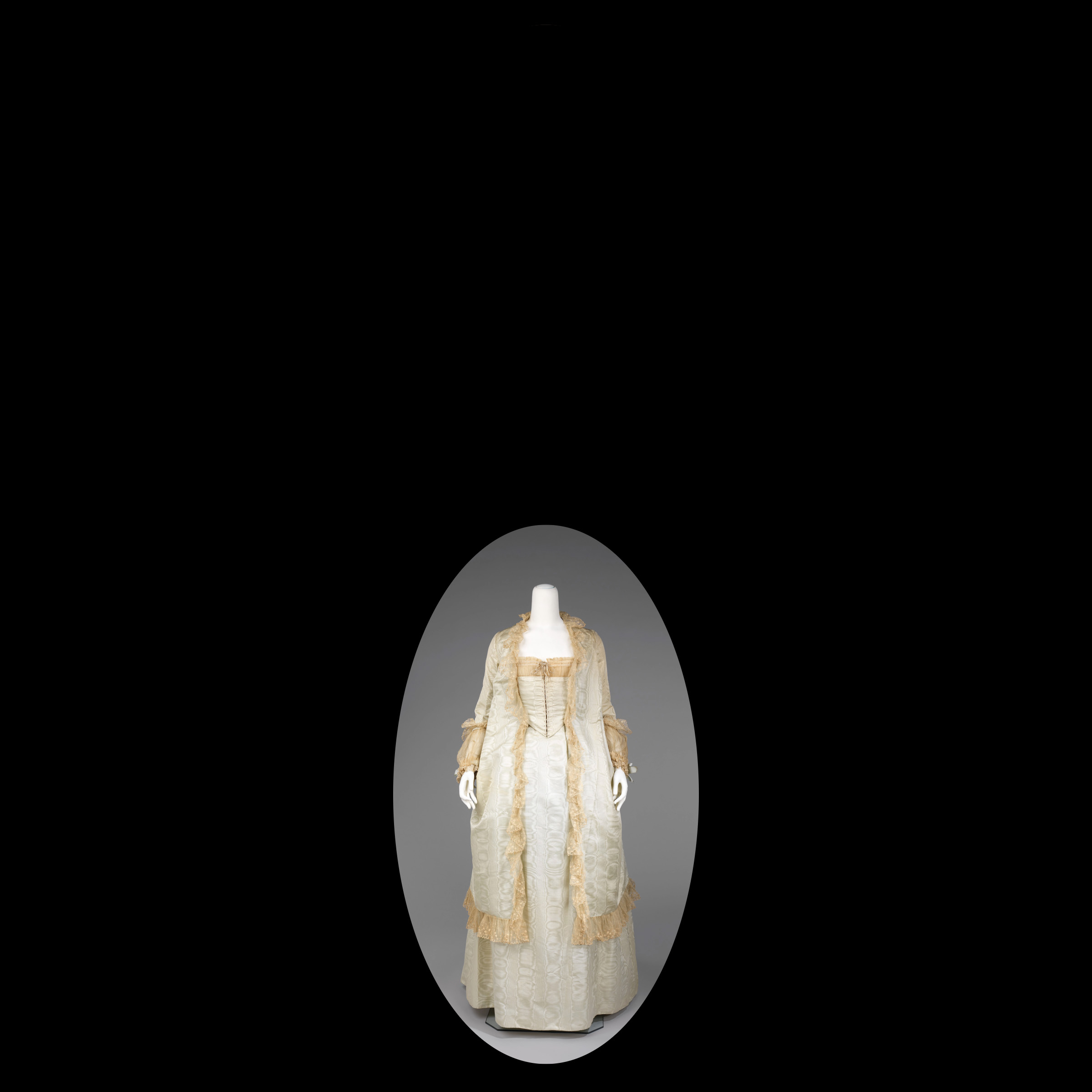 Oval crop of a 19th-century tea gown, made of white and gold fabrics and materials, with a black background.