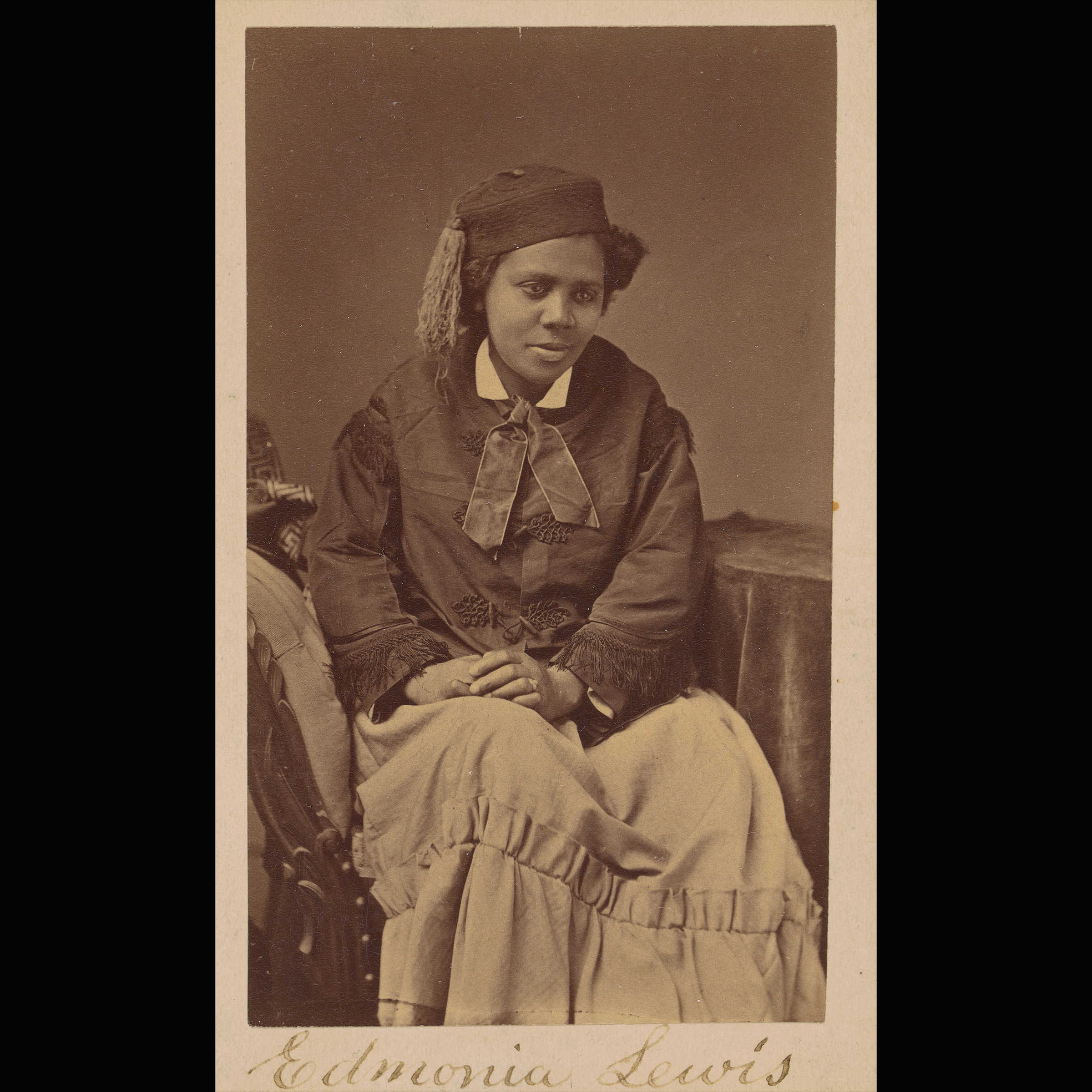 Archival photograph of Black woman seated, facing the camera, wearing a hat, jacket, and dress.