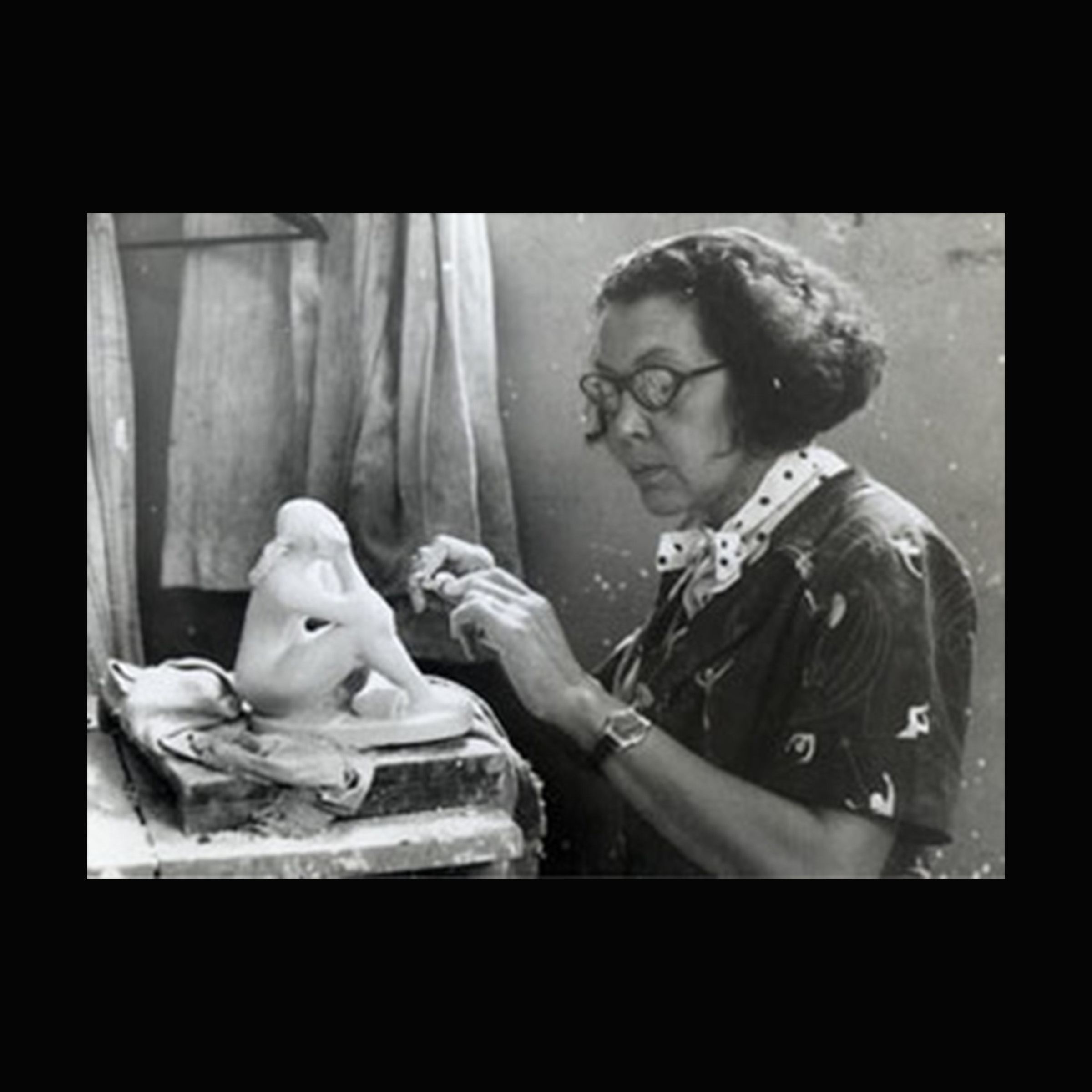 A seated Black woman artist with both hands raised toward a sculpture in progress.